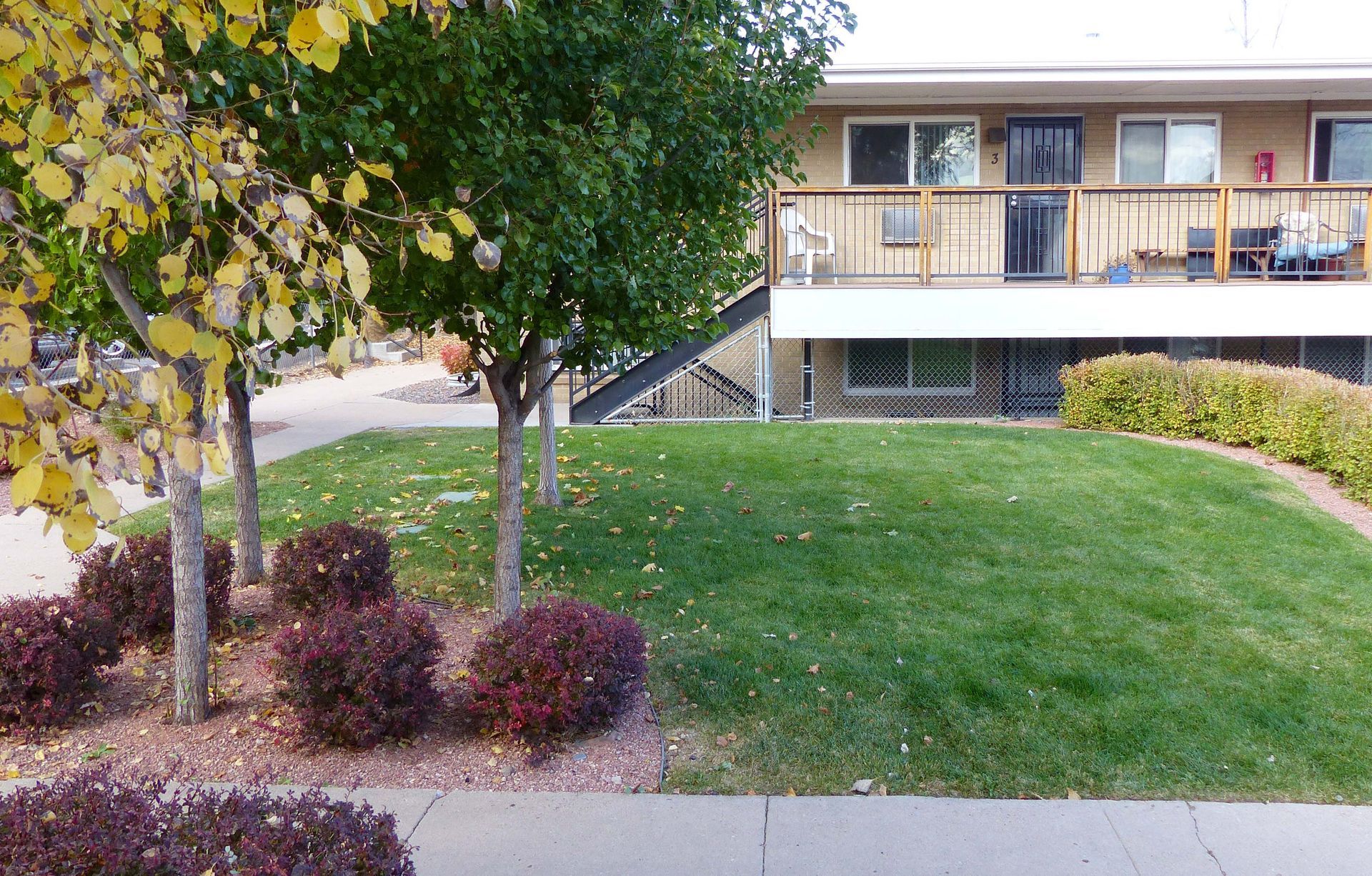 Apartment building beside a green lawn with trees and a sidewalk in the foreground