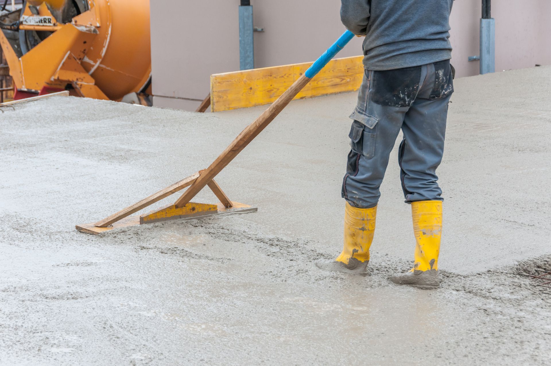 Person smoothing wet concrete with a screed tool, wearing yellow boots and gray pants, at a construction site.