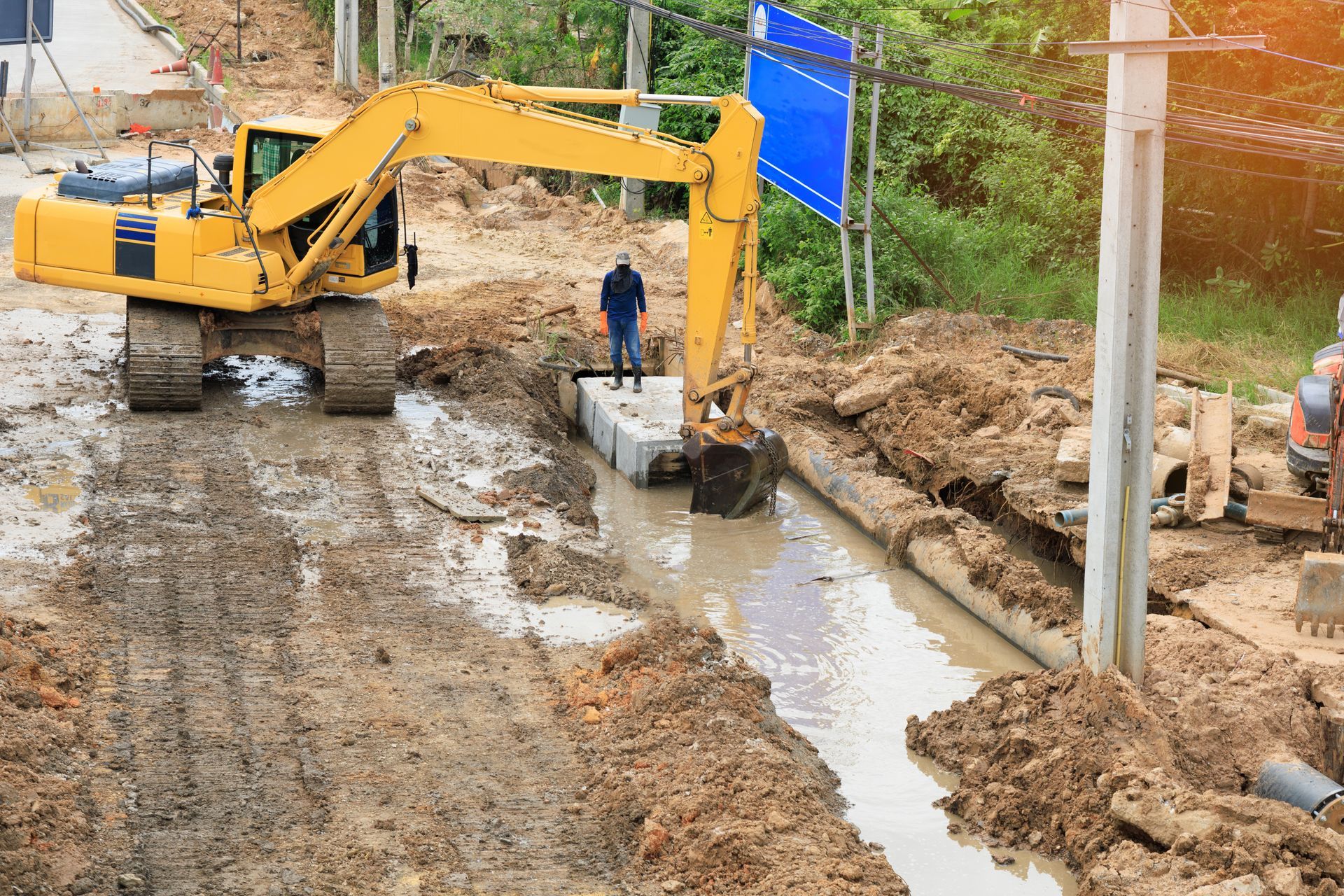 Yellow excavator working at a construction site in mud; a person stands nearby.