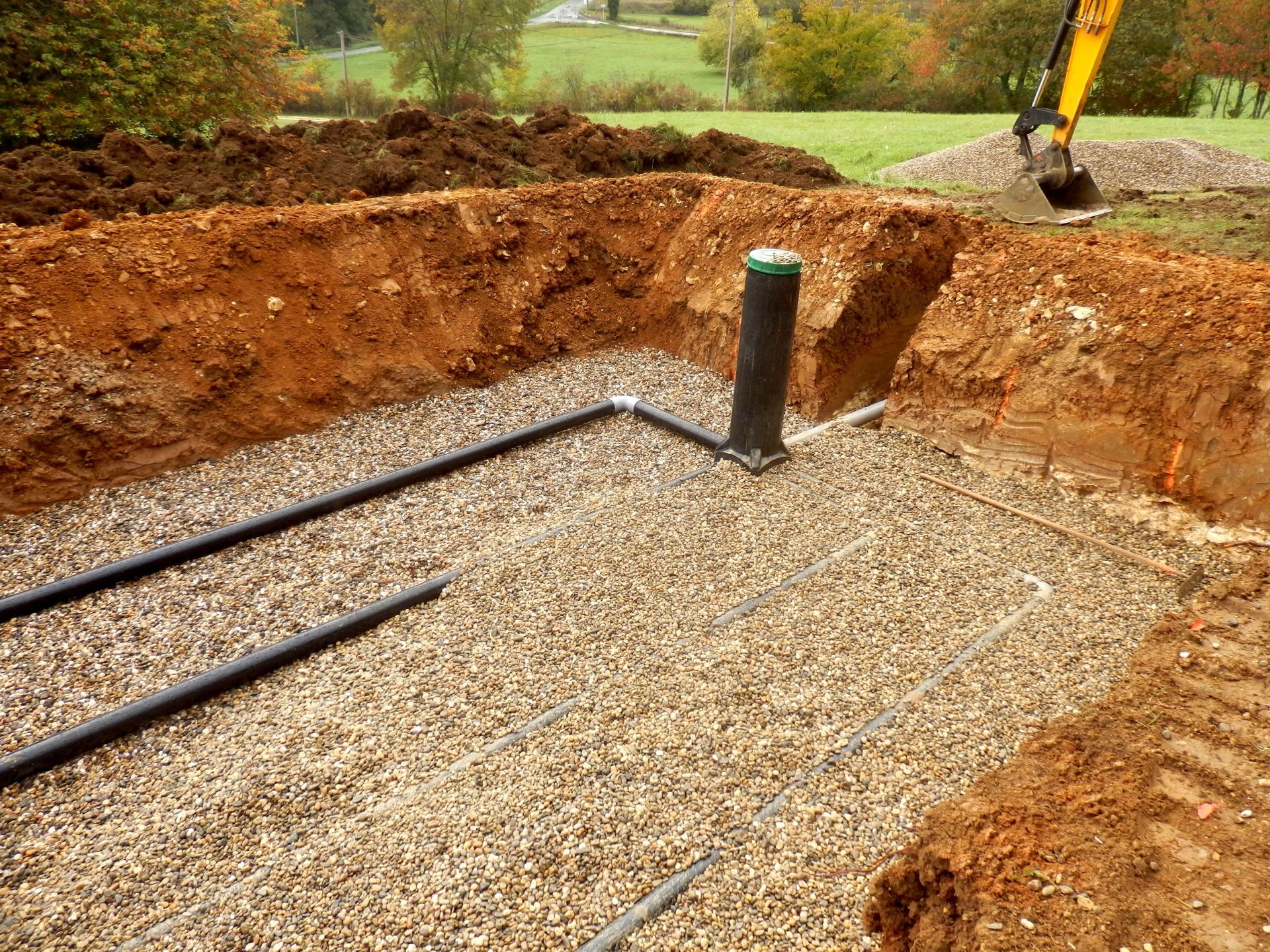 Excavation pit with gravel, black pipes, and a vertical pipe; construction site outdoors.