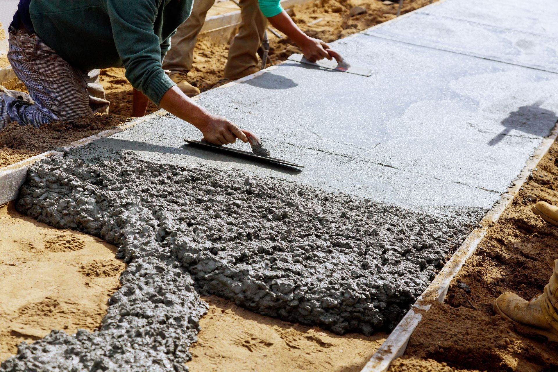 Workers smoothing wet concrete for a sidewalk with trowels, wooden frame set in sandy ground.
