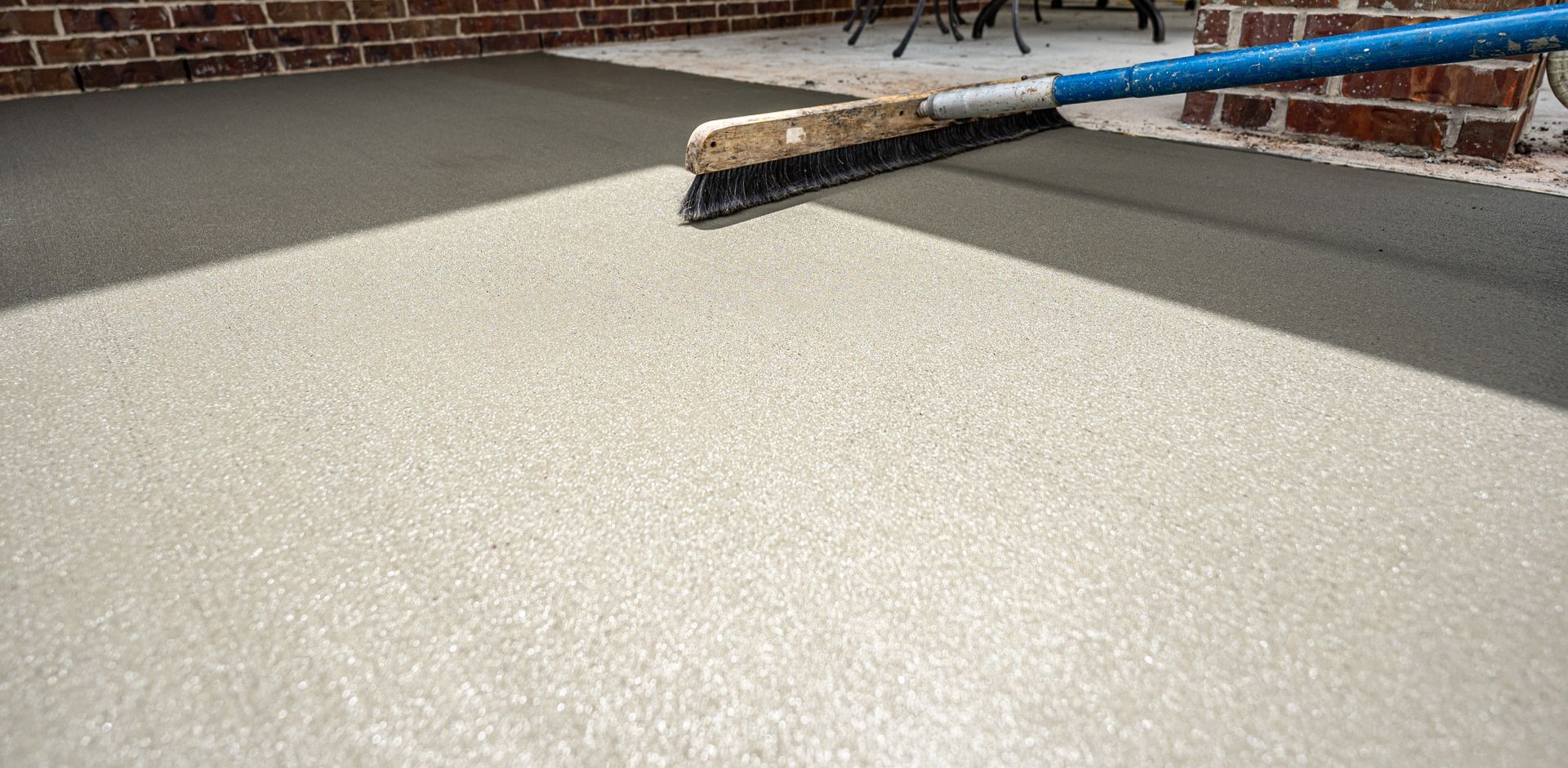 A person using a broom to spread a light gray concrete surface on a patio. Brick wall is visible.