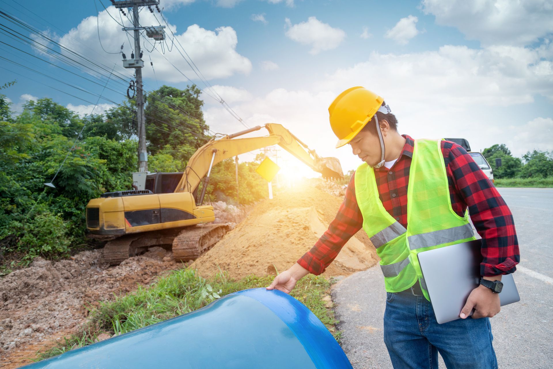 Construction worker inspecting blue pipe near excavator on roadside. Sunny day.