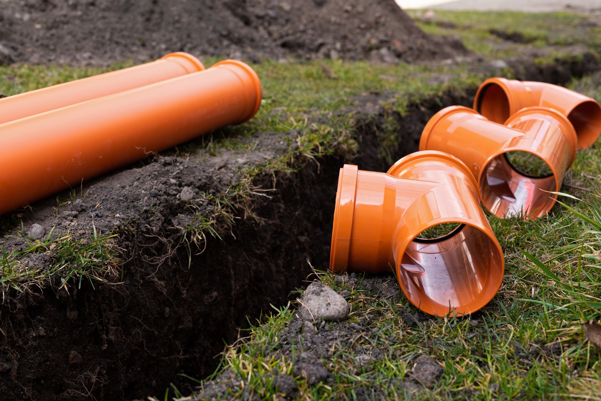 Orange PVC pipes and elbows on grass near a ditch, ready for installation.
