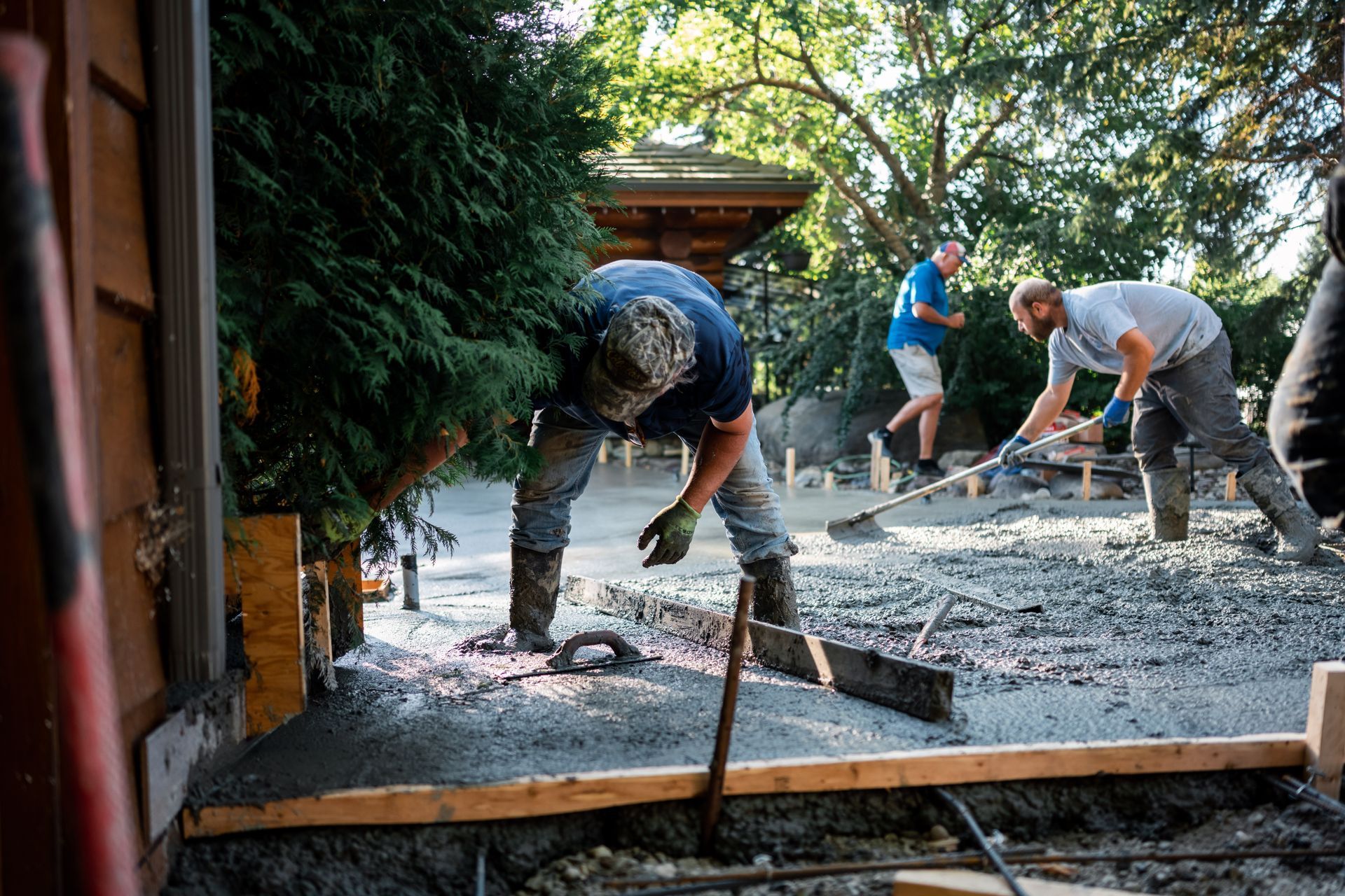 Workers laying concrete; one bending over, two others shoveling in a sunny outdoor setting.