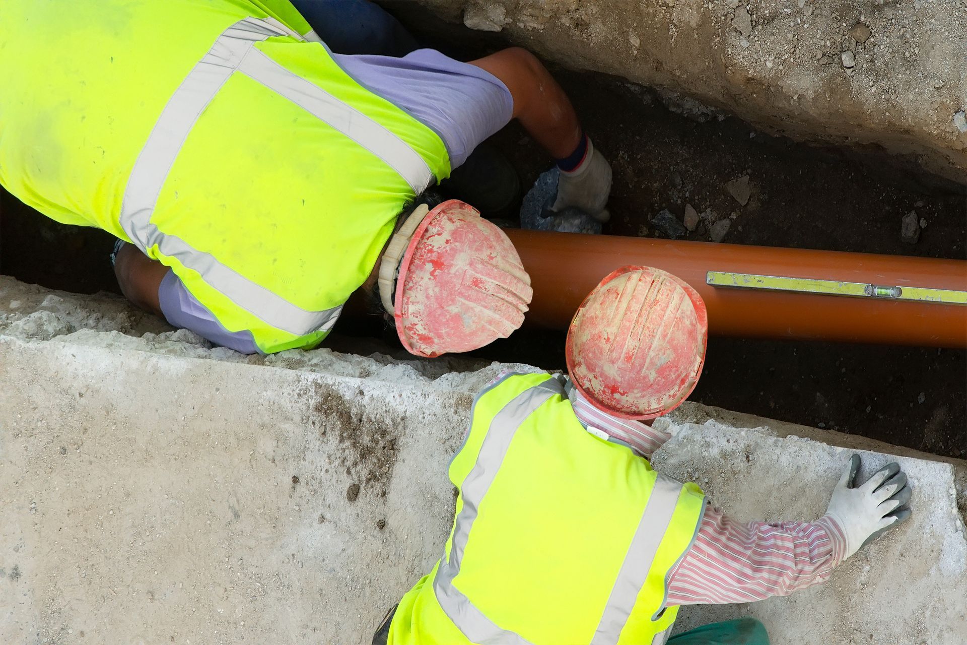 Two construction workers installing a brown pipe in a trench, wearing safety vests and hard hats.