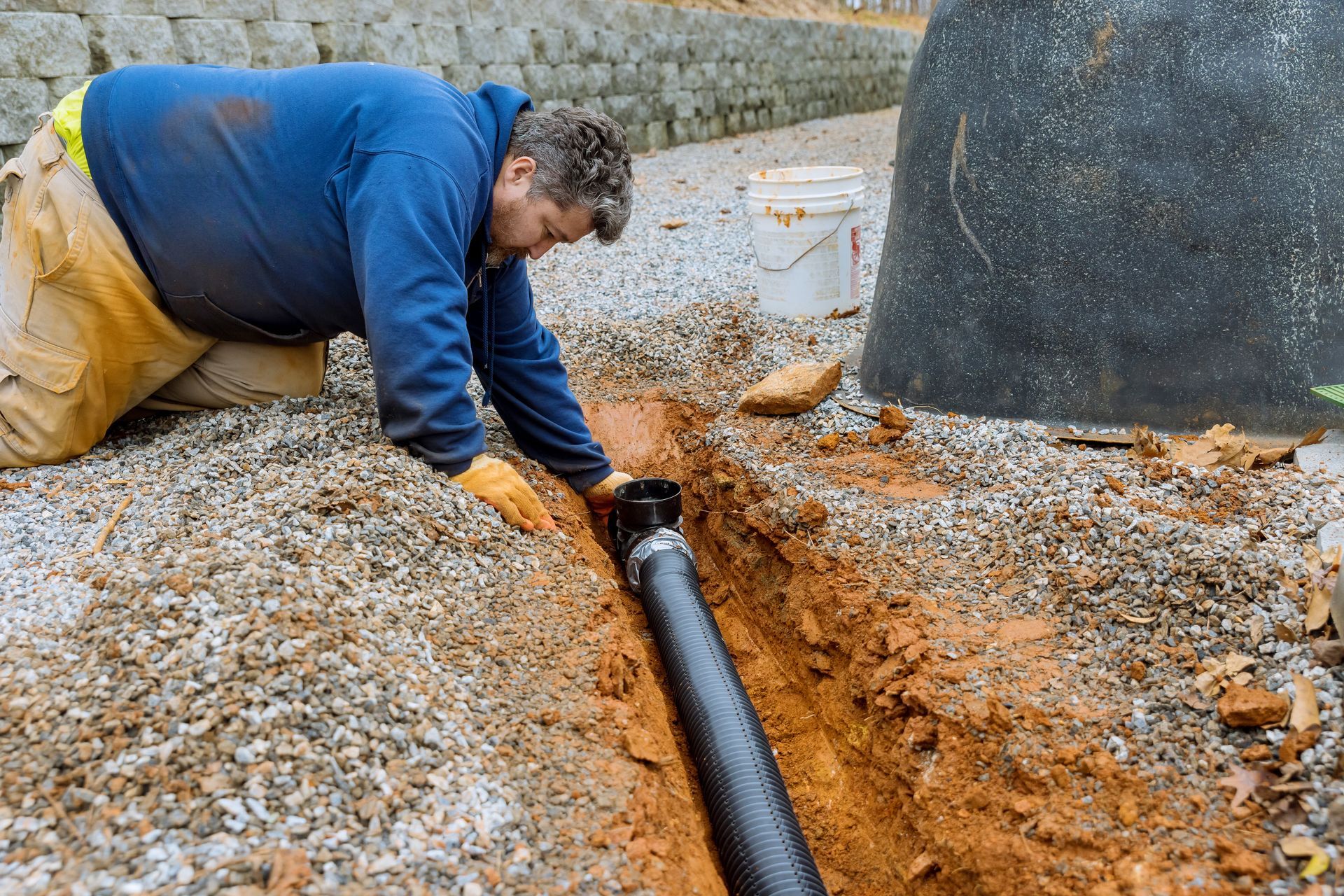 Man installing black drainage pipe in a gravel-lined trench outdoors.