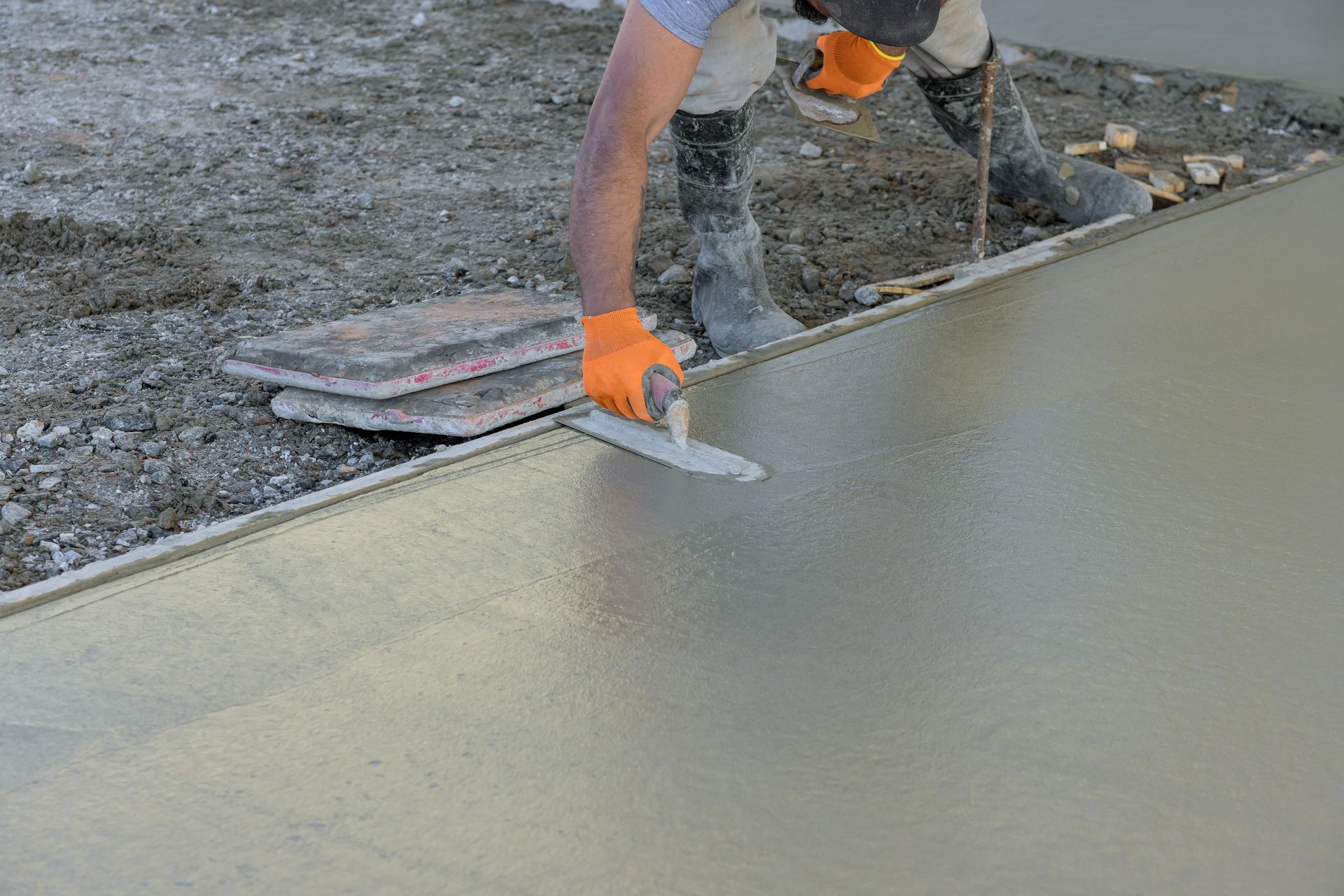 Person in boots and gloves smoothing wet concrete with a trowel outdoors.