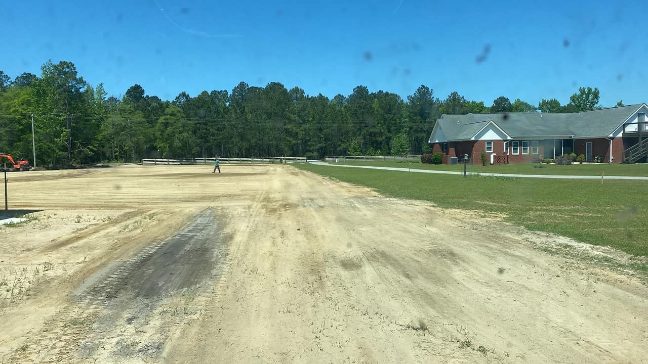 A leveled, sandy construction lot next to a brick building, with a forest in the background under a clear blue sky.