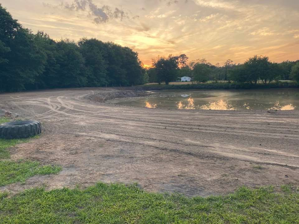 A dirt-covered shoreline by a pond at sunset, with tire tracks in the mud and trees in the background.