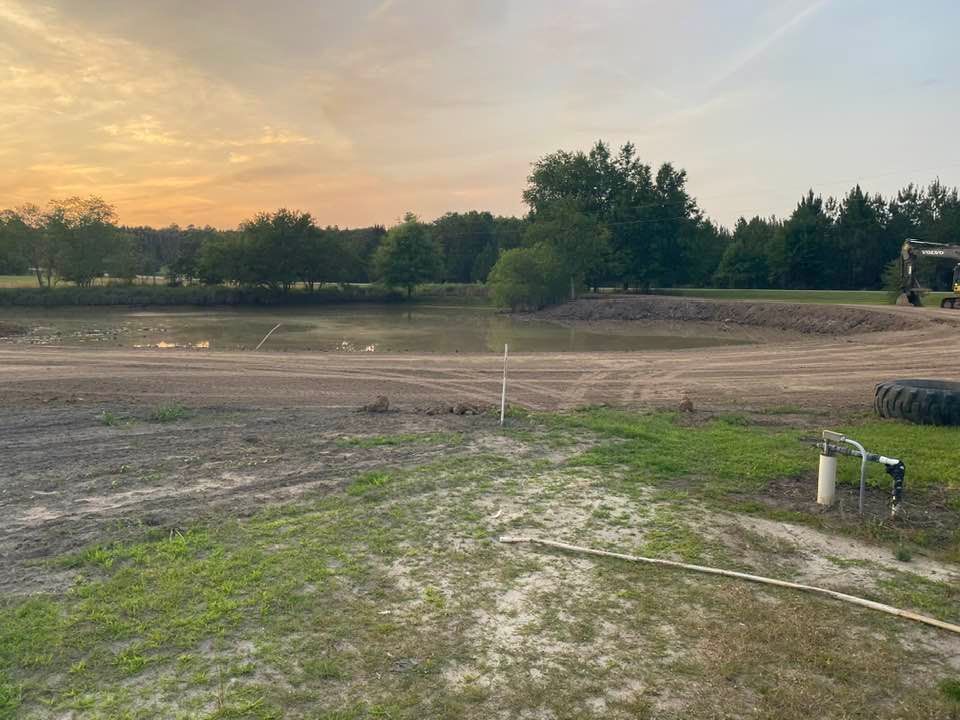 A muddy pond at sunset with cleared land in the foreground, a few trees in the distance, and heavy machinery parked nearby.