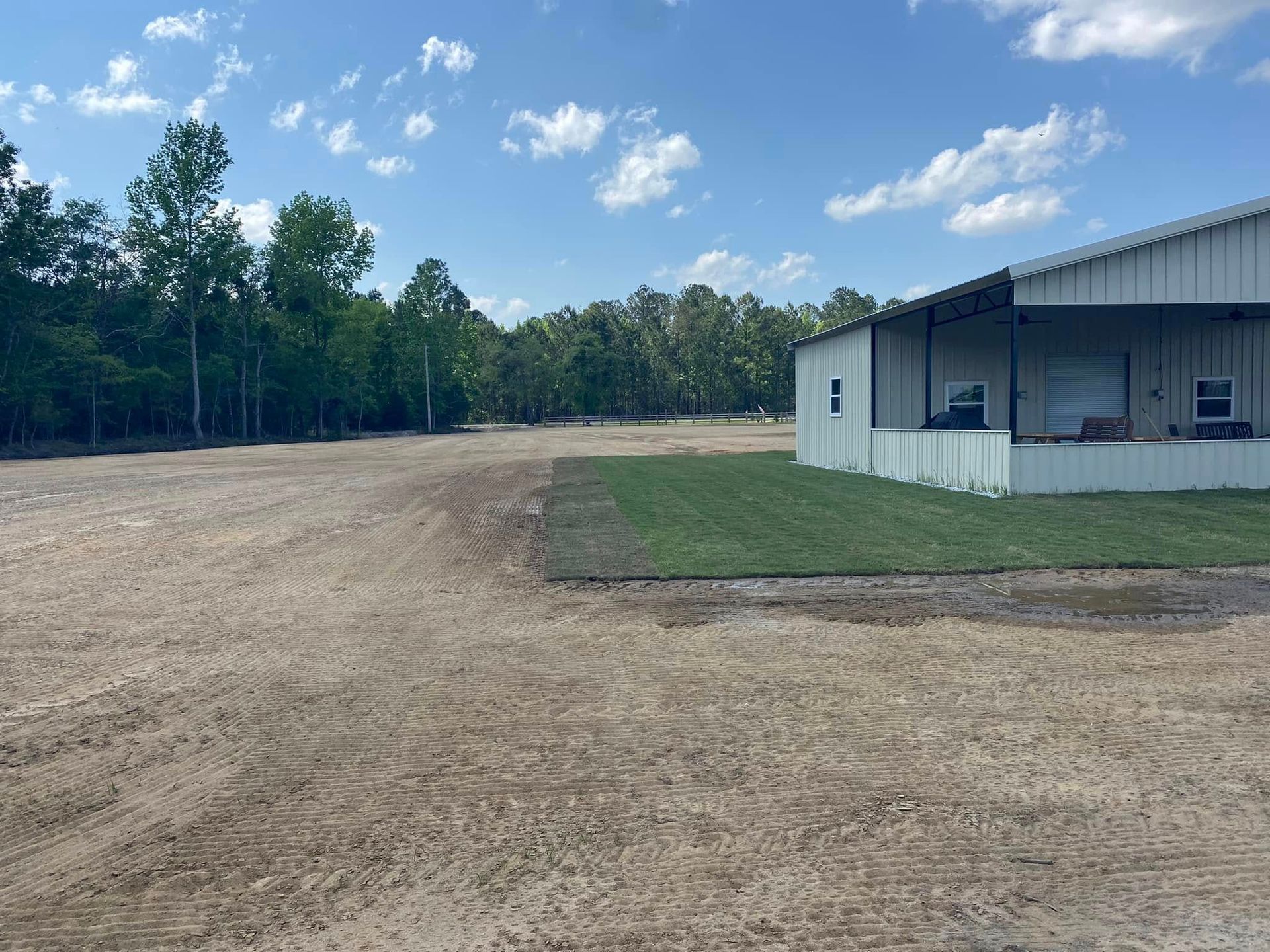 A white metal building sits beside a patch of green grass, overlooking a large, cleared dirt lot lined by tall trees.