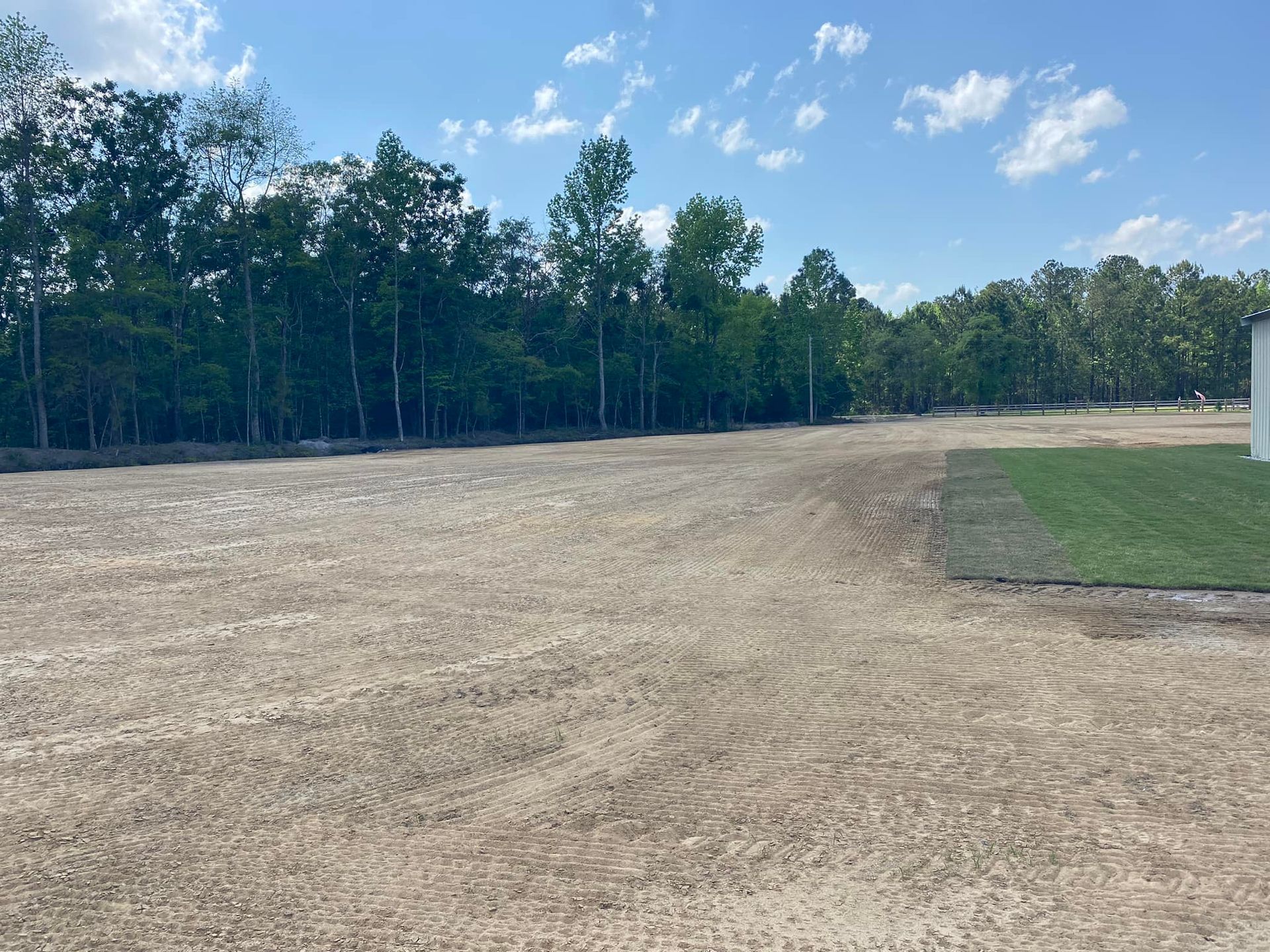 A large, cleared construction site with a freshly sodded green lawn edge next to a dense line of green trees.
