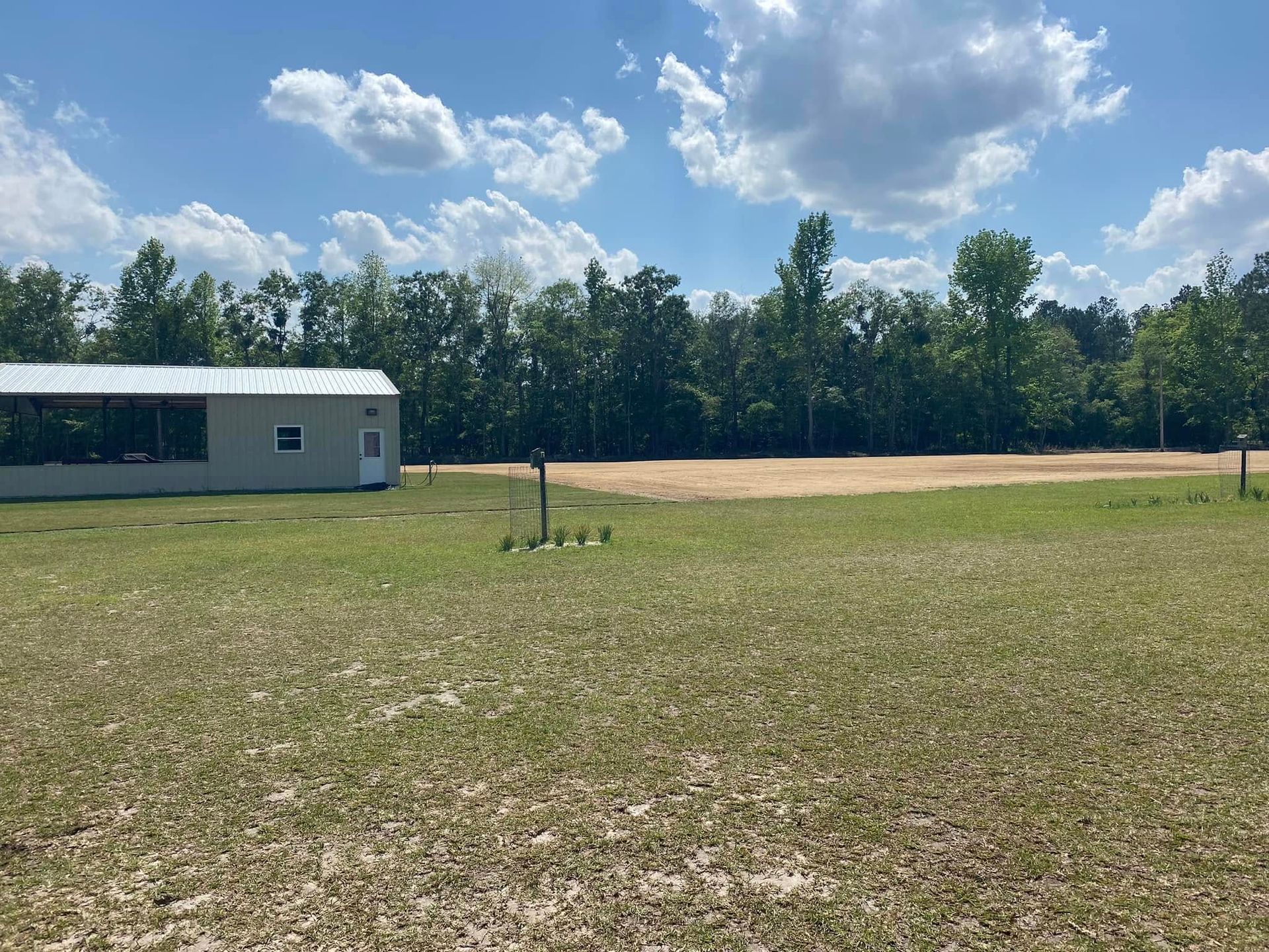 A light-colored metal building sits on a grassy lot next to a cleared, sandy area with a forest line in the background.