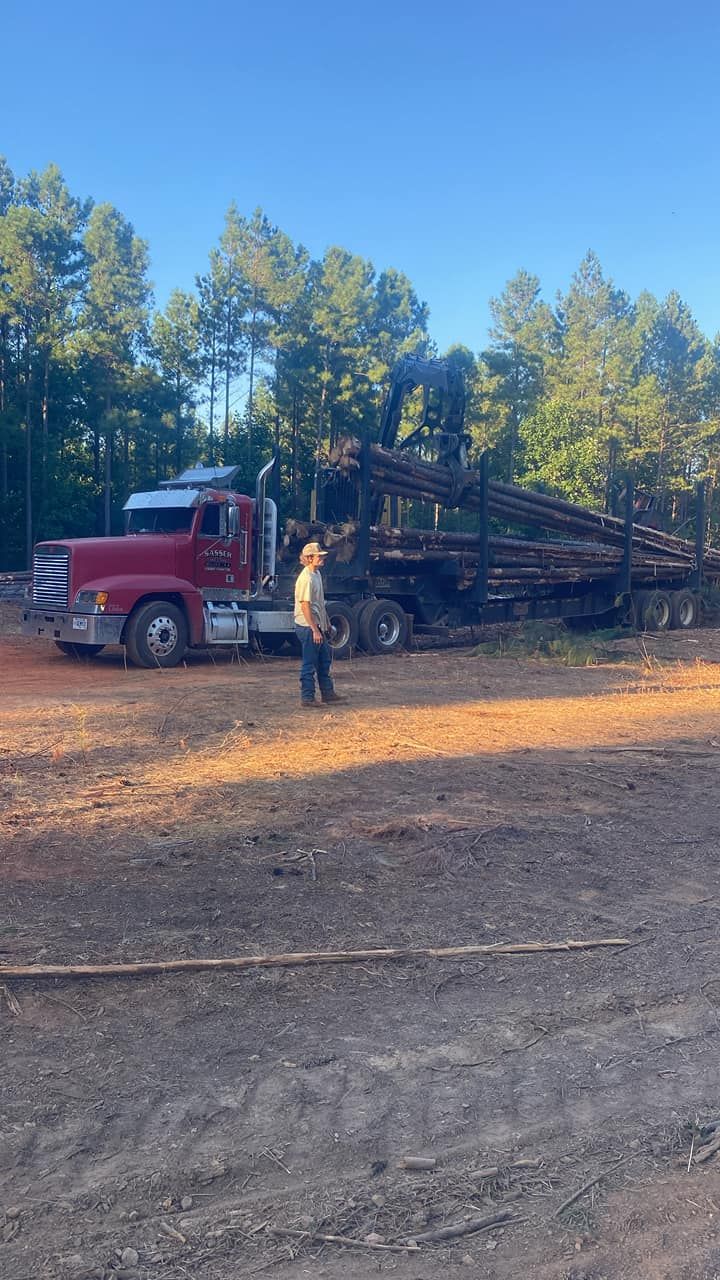 A person stands beside a red logging truck being loaded with timber in a forest clearing on a sunny day.