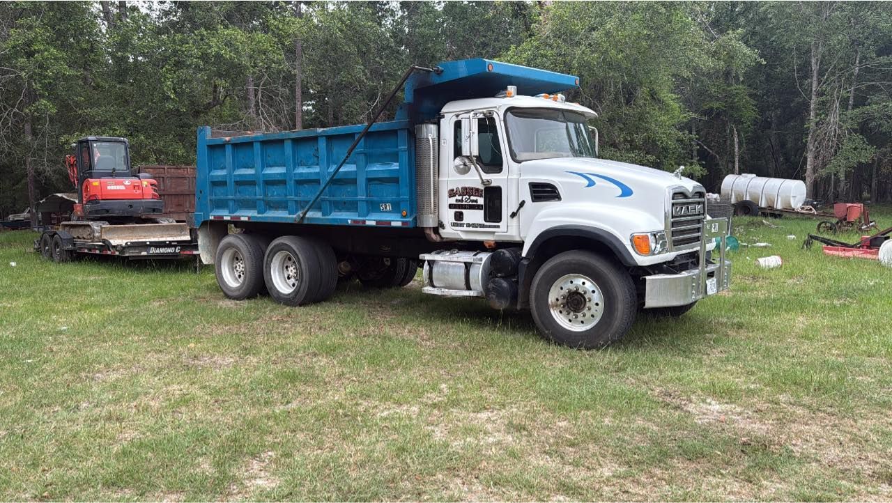 A white Mack dump truck with a blue bed is parked on a grassy lot, pulling a trailer carrying a small orange excavator.