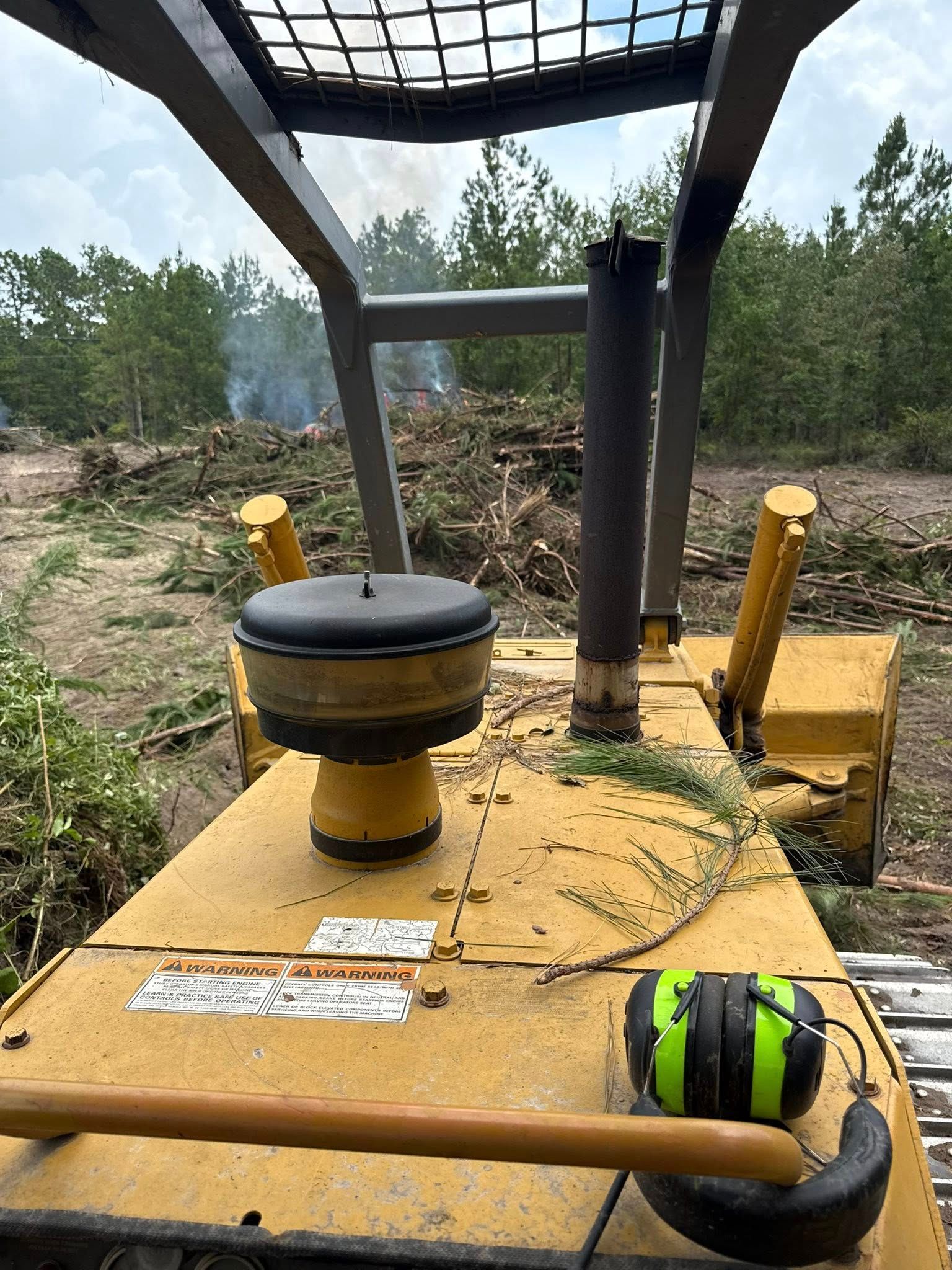 View from inside a yellow bulldozer looking out at a forest clearing with smoke in the distance.