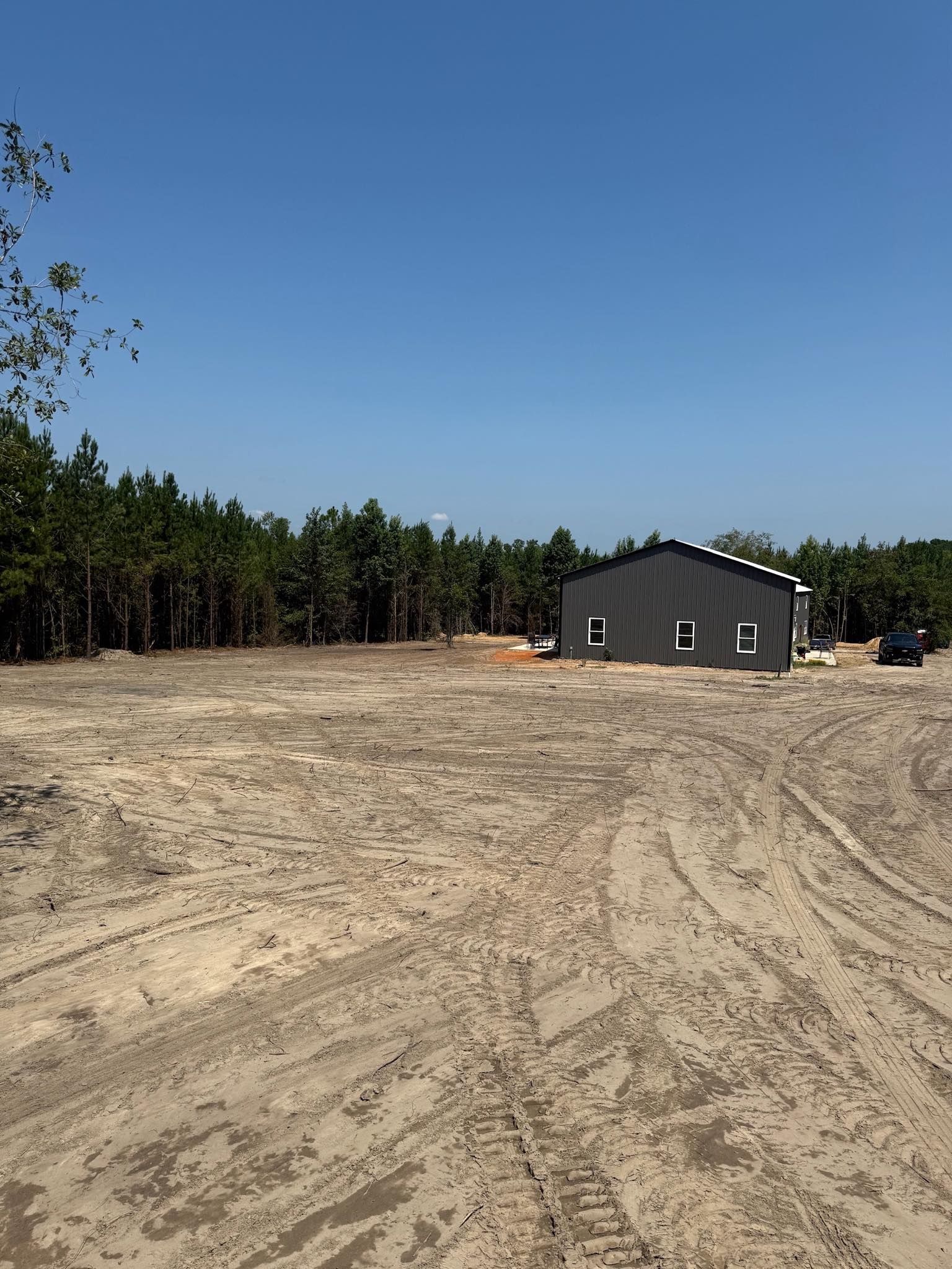 A dark, modern, single-story house sits on a large, cleared dirt lot in front of a dense pine forest under a blue sky.