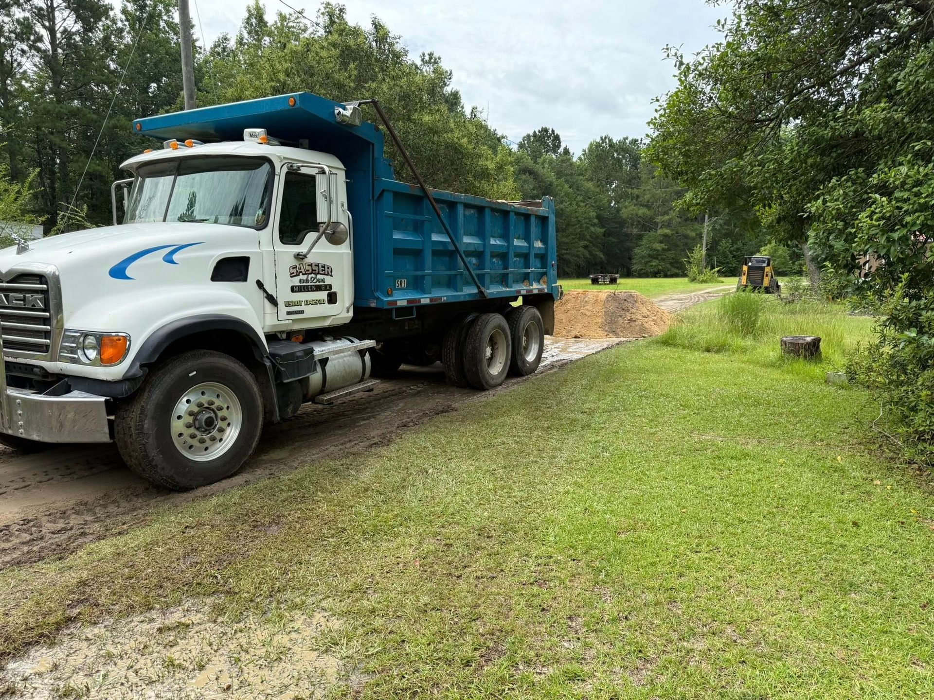 A white dump truck with a blue cargo bed is parked on a dirt path next to a grassy field with a small pile of dirt.