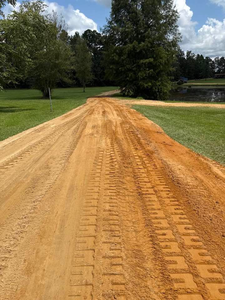A freshly graded dirt road with deep tire tracks, surrounded by green grass, trees, and a small pond under a blue sky.