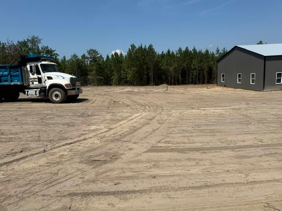 A white dump truck sits on a large, cleared dirt lot next to a gray metal building in front of a dense pine forest.