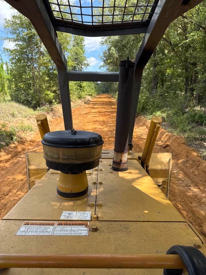 View from inside a bulldozer cab looking out over a freshly cleared red dirt path through a forest.