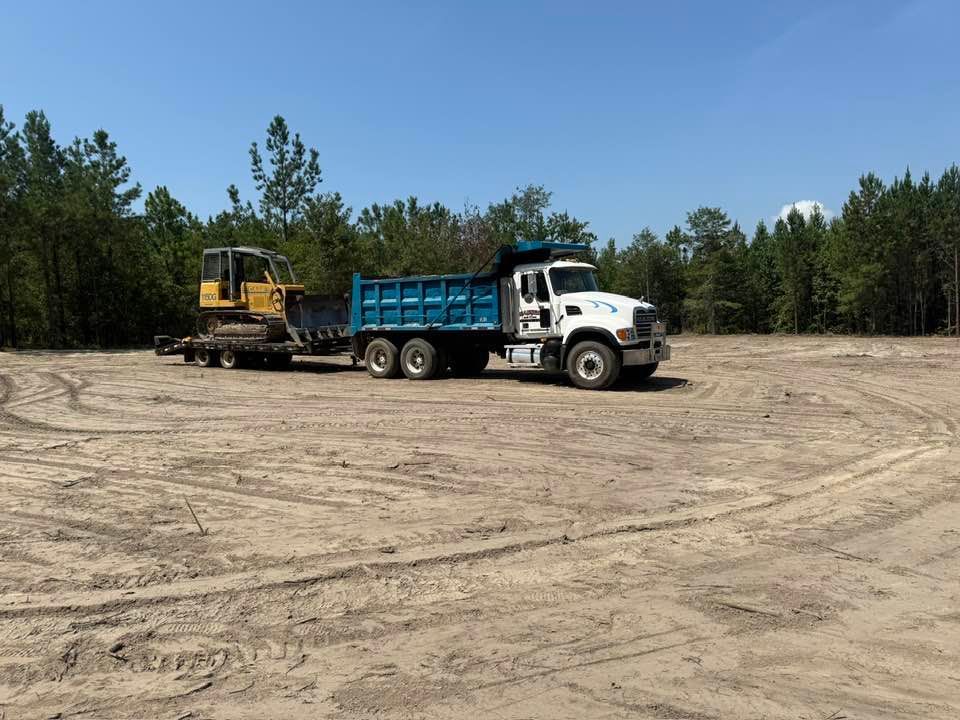 A white dump truck is hauling a yellow bulldozer on a flatbed trailer across a cleared, sandy construction site.
