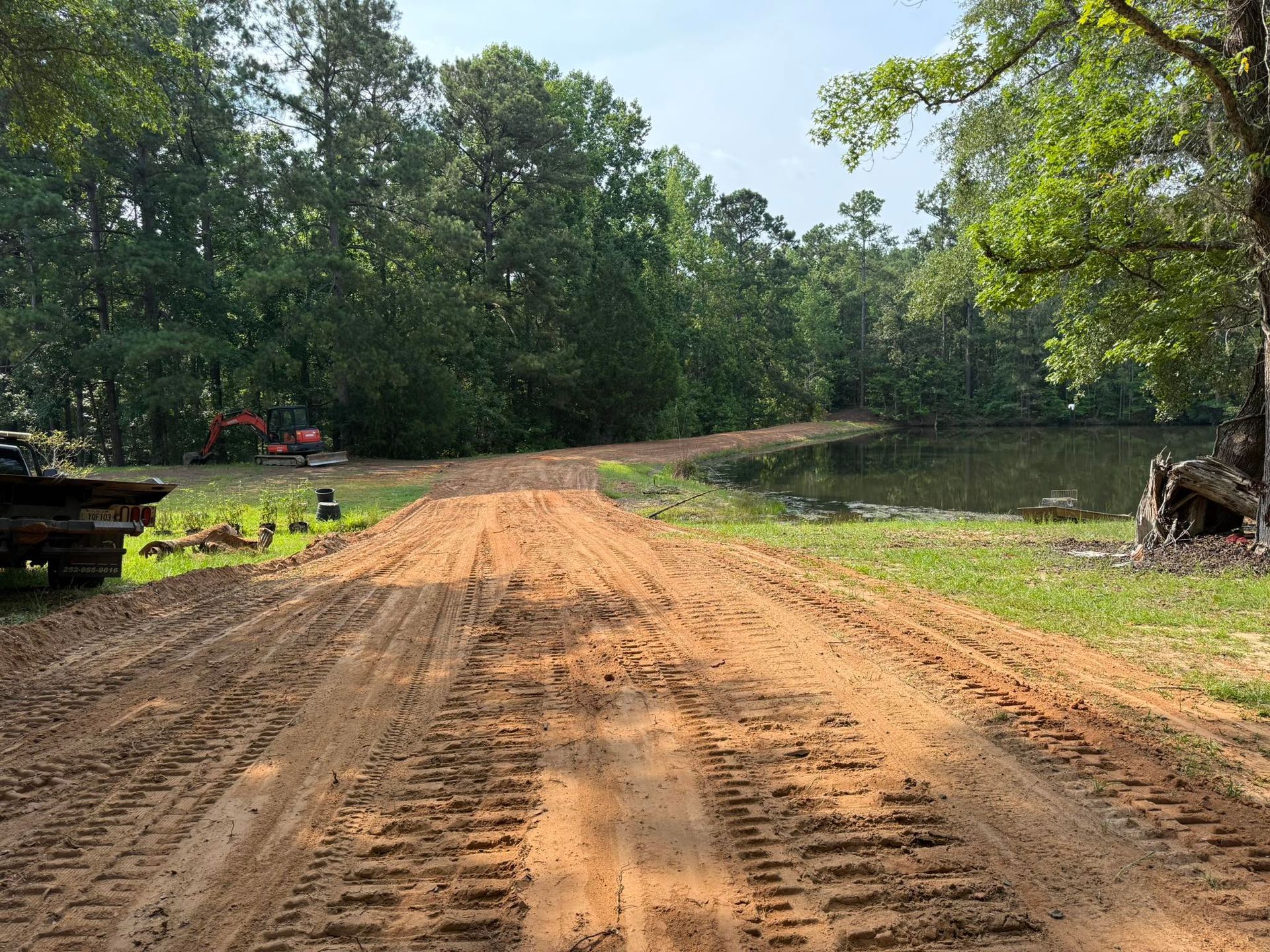 A dirt road with tire tracks leads toward a pond bordered by trees, with heavy construction machinery visible nearby.