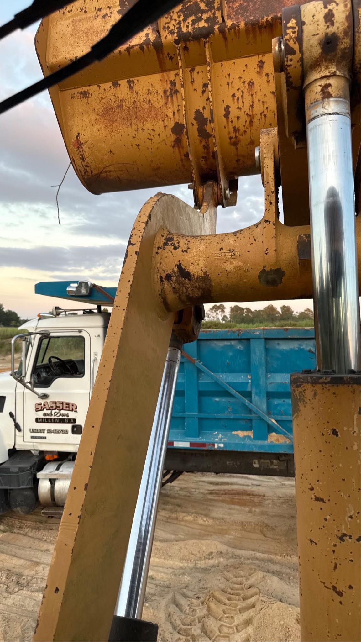 A close-up view from an excavator cab showing the yellow bucket and hydraulic lift, with a truck parked in the background.