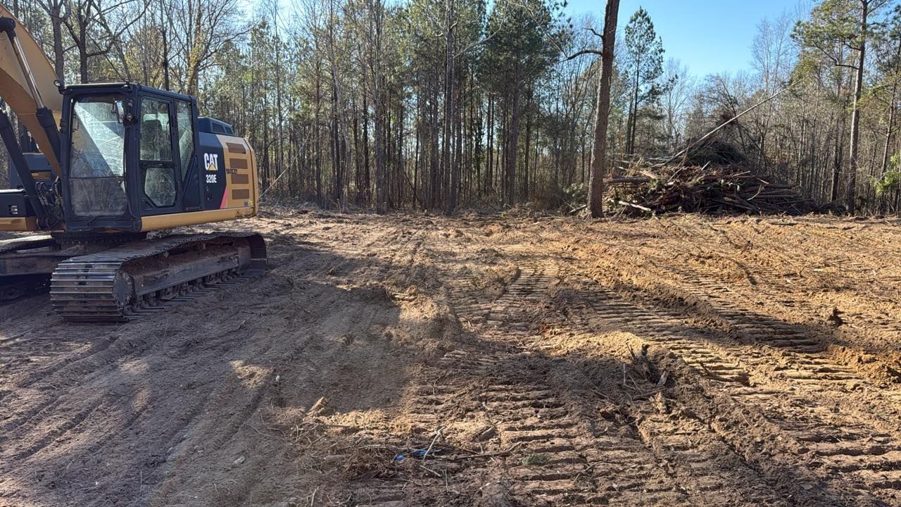 A yellow Caterpillar excavator sits on a dirt lot, near a cleared forest area with a pile of wood debris.