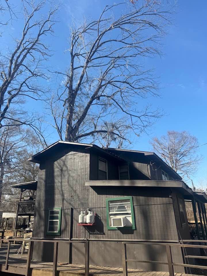 A dark, two-story wooden cabin with a wraparound deck, set against a backdrop of bare trees under a clear blue sky.