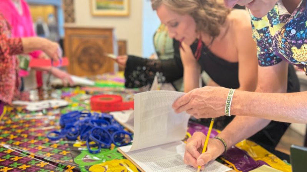 People gather around a table covered with craft supplies, one person writes in a notebook with a yellow pencil.