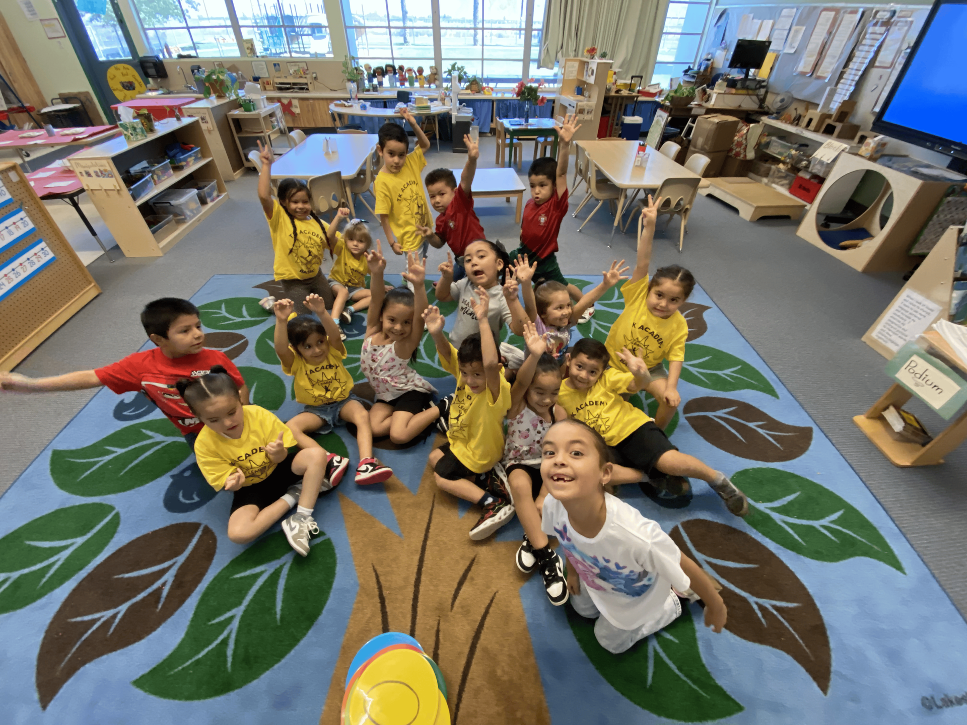 An adult sits on a blue rug and points to a colorful, interactive toy as a young child engages with it.