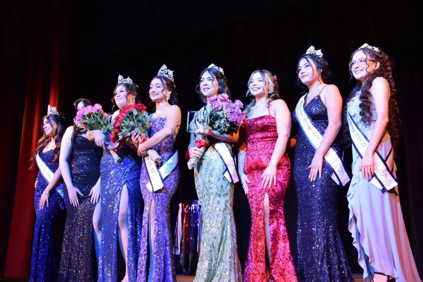 A woman speaks into a microphone while others wearing sashes and crowns stand behind seated guests at a formal event.