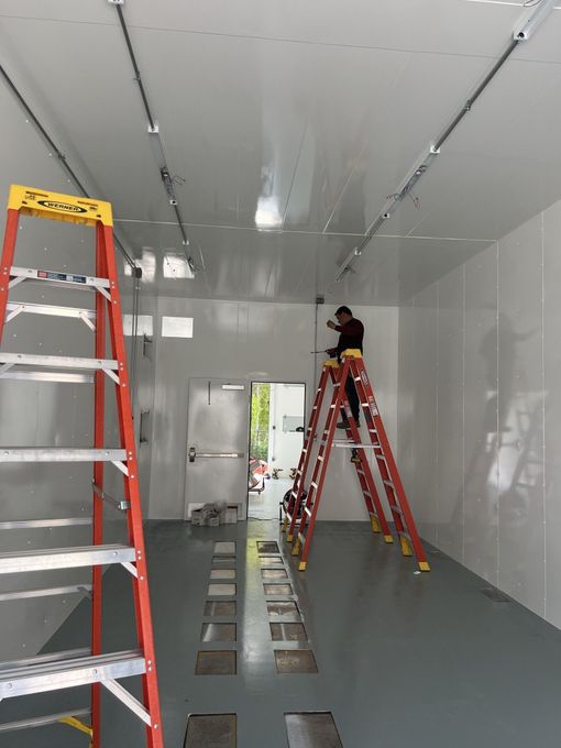 Person on a ladder installs ceiling fixtures in a white-walled room with a gray floor.