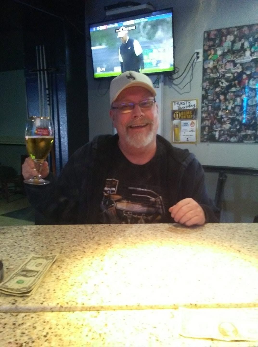Man smiling at a bar, holding up a drink, with a TV and wall art behind him.