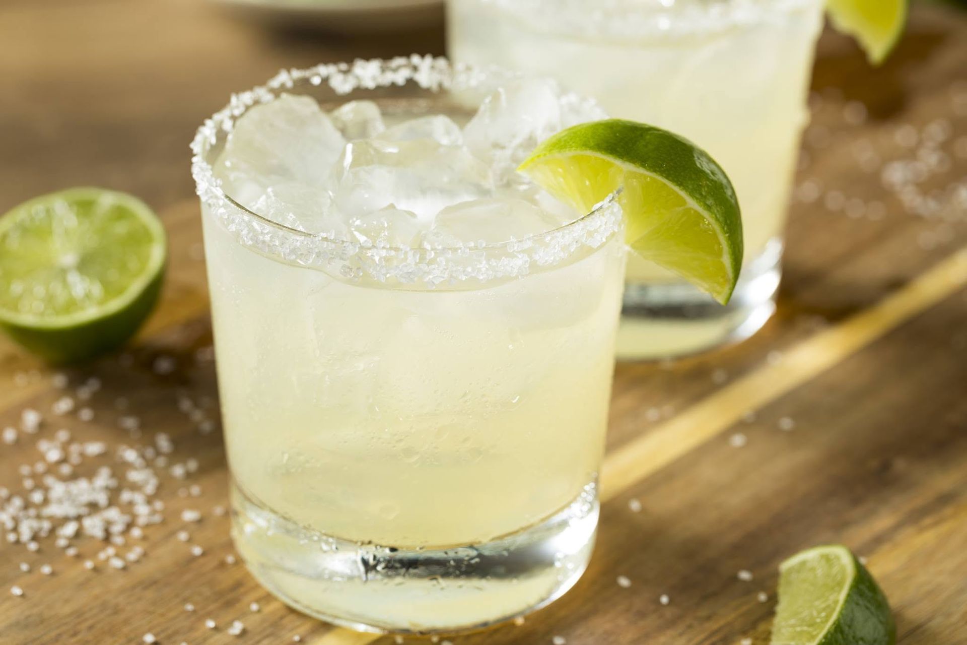 Two margarita glasses with lime wedges and salt rims on a wooden table, with halved limes nearby
