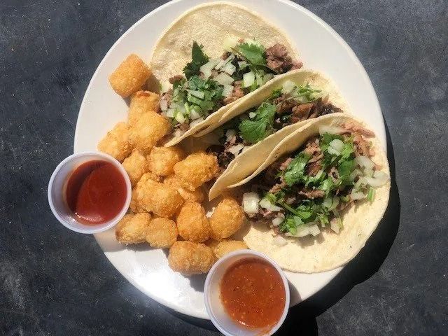 Tacos with cilantro and onions on a plate, with tater tots and two small cups of red sauce.