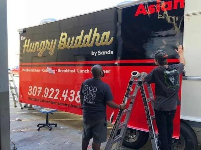 Two workers applying lettering to a red-and-black food truck wrapped with “The Hungry Buddha” branding.