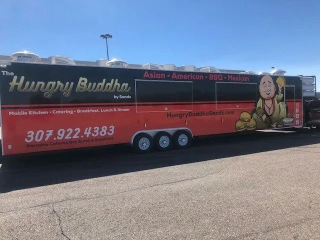Red Hungry Buddha food truck parked in a lot under a blue sky, with menu graphics and phone number displayed