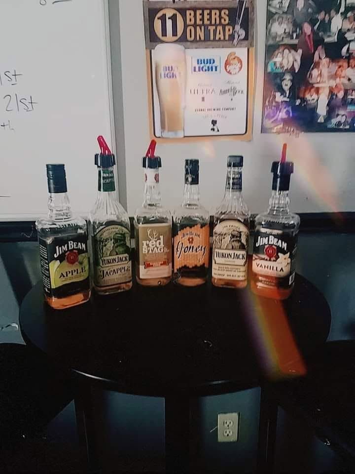Six liquor bottles lined up on a black table beneath a “Beers on Tap” sign.