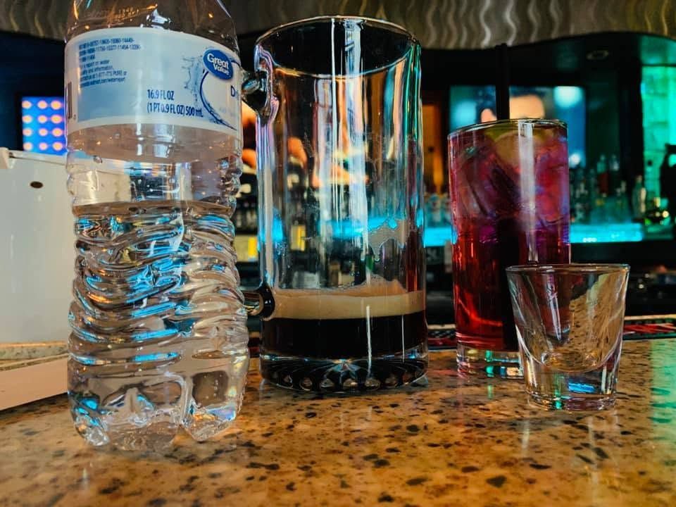 Water bottle and cocktails on a bar counter with a small shot glass.