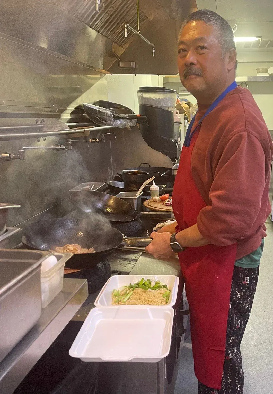Chef in red apron cooking at a steaming stove in a busy kitchen