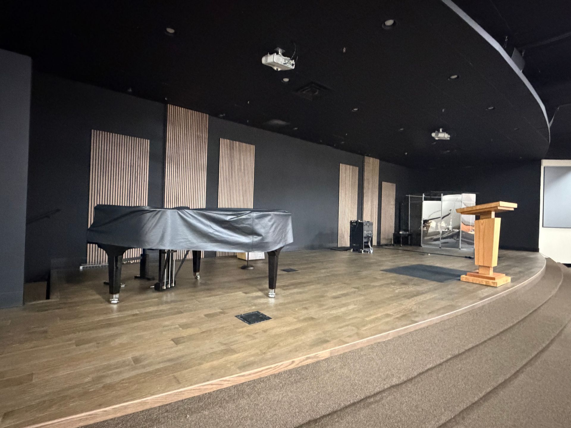 Construction in a black-walled room with a stage. Covered equipment, a worker, and exposed drywall are visible.