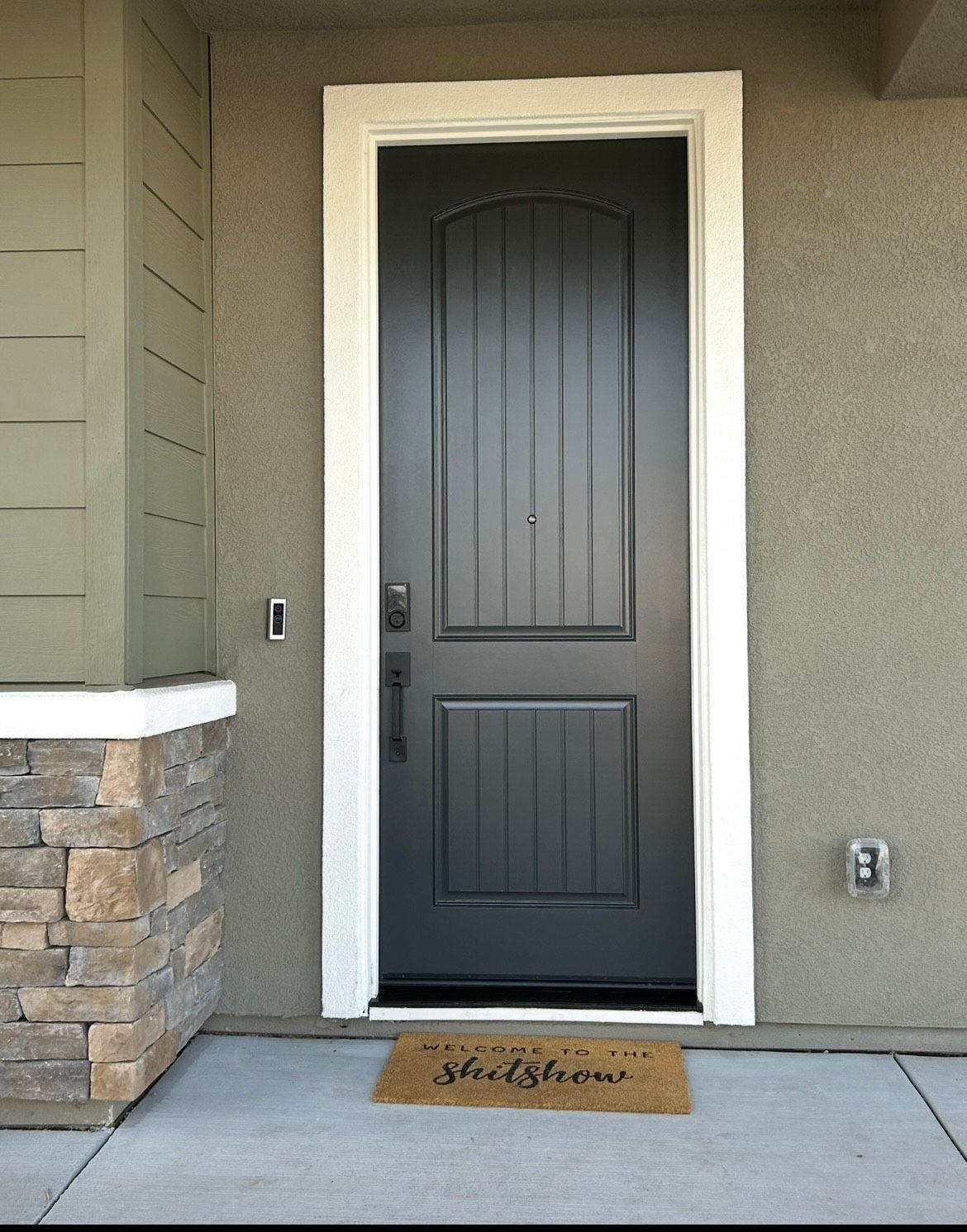 Black front door with white trim, tan welcome mat, and stucco exterior.