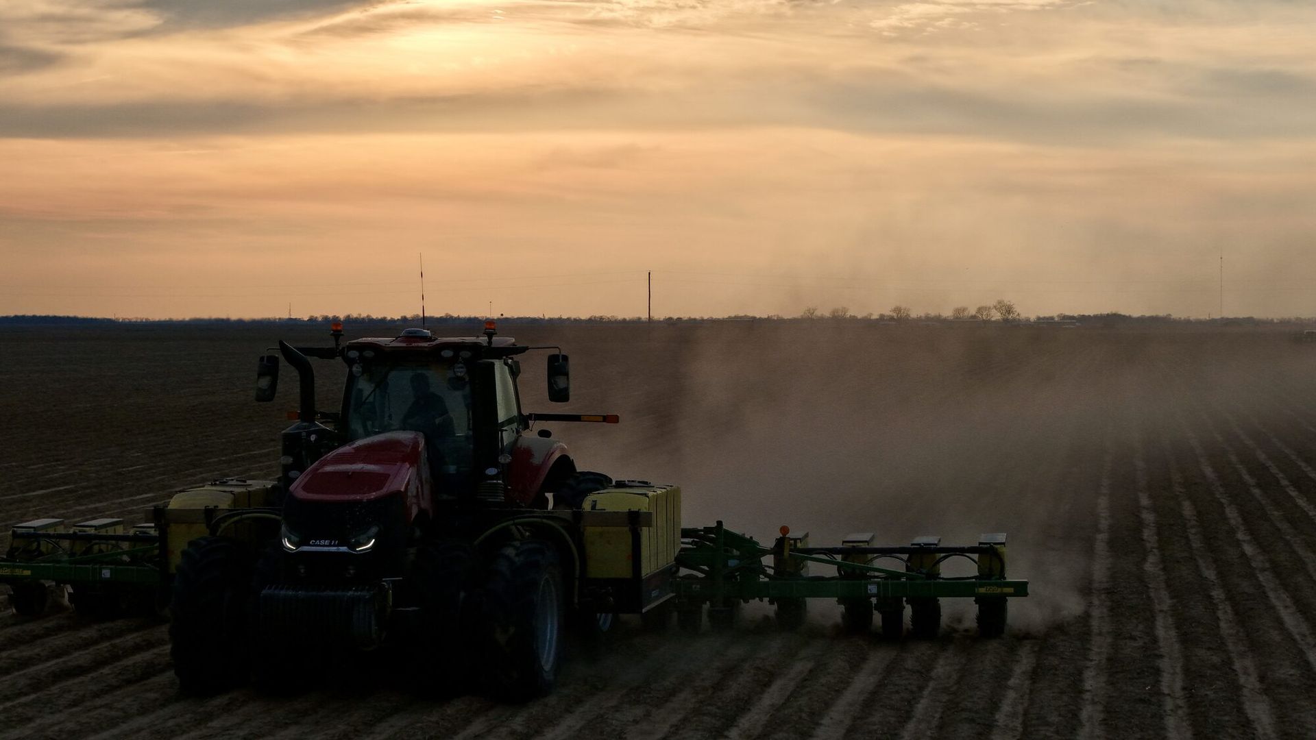 Tractor plowing a field at sunset, kicking up dust under a cloudy sky
