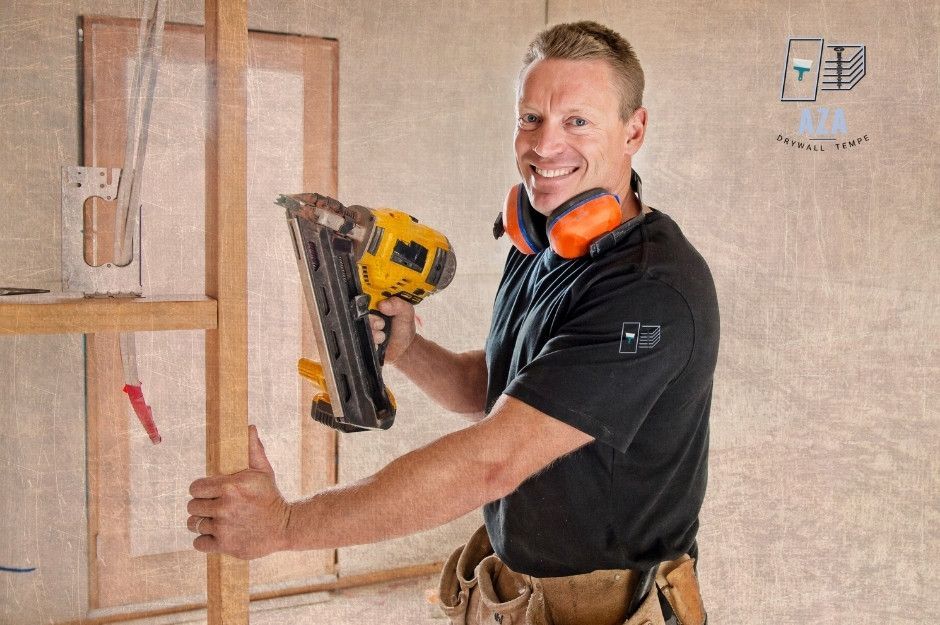 A drywall repair company worker smiles while using a pneumatic nail gun on wooden framing. He is wearing a black shirt, tool belt, and safety gear in an unfinished residential construction area with exposed studs and drywall sheets near W Grand Canyon Dr, and S Dobson Rd, Chandler, AZ.