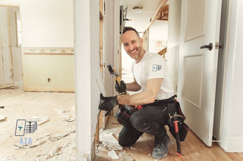 A drywall repair company professional kneeling on plastic sheeting, holding a power tool while working on wall repairs in a residential hallway. The worker wears a white shirt and dark pants, with tools nearby near N Four Peaks Way, and E Firerock Country Club Dr, Fountain Hills, AZ.