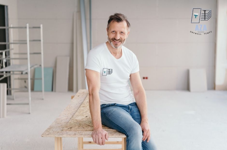 A drywall repair company professional in a white t-shirt and jeans sits on stacked drywall sheets in a bedroom under a residential renovation. The room has exposed walls and flooring materials visible in the background near E Twin Acrs Ct, and E 214th Way, Queen Creek, AZ.