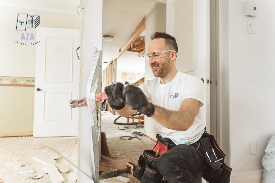A drywall repair company professional wearing a white t-shirt and tool belt kneels while using a power tool on white drywall. He is working in a residential room with wooden flooring and white walls during renovation near E Plover Ct, and Forest Rd, Rio Verde, AZ.